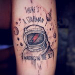 Фото тату на предплечье с изображением космического шлема и надписью 'There's a Starman waiting in the sky'. Черно-серый рисунок татуировки в стиле графика.