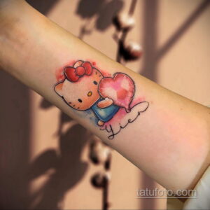 Цветное фото татуировки с изображением Hello Kitty, держащей красное сердце. Под сердцем надпись "Lien". Рисунок выполнен в акварельном стиле.