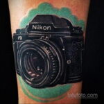 Фото тату реалистичного фотоаппарата Nikon, выполненного в черно-серых тонах с акварельным зеленым фоном. Детализированный рисунок татуировки.