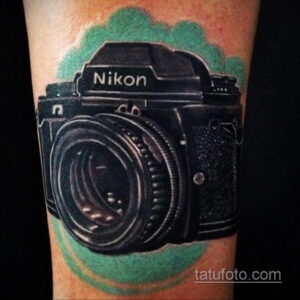 Фото тату реалистичного фотоаппарата Nikon, выполненного в черно-серых тонах с акварельным зеленым фоном. Детализированный рисунок татуировки.