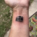 Фото тату маленького черного фотоаппарата на запястье. Изображение выполнено в минималистичном стиле, с четкими контурами.