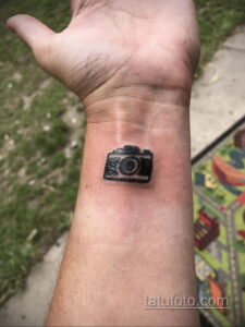 Фото тату маленького черного фотоаппарата на запястье. Изображение выполнено в минималистичном стиле, с четкими контурами.