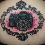 Фото тату фотоаппарата Canon, обрамленного винтажной рамкой с розовыми акцентами, выполненное в черно-серых тонах на спине.
