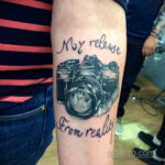 Фото тату винтажного фотоаппарата с надписью 'My release from reality' на руке, выполненное в стиле графика с использованием черных чернил.