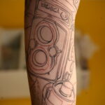 Детальный эскиз татуировки старинного фотоаппарата Yashica на руке. Рисунок выполнен тонкими черными линиями с добавлением красных акцентов.