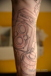 Детальный эскиз татуировки старинного фотоаппарата Yashica на руке. Рисунок выполнен тонкими черными линиями с добавлением красных акцентов.