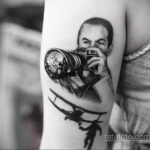 Черно-белое фото тату на предплечье с изображением фотографа с камерой и силуэтом самолета внизу. Детализированный рисунок, реалистичный стиль.