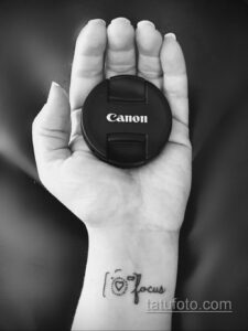 Фотография татуировки на запястье с изображением объектива Canon и надписи 'Focus'. Черно-белый рисунок с четкими линиями.