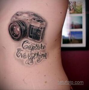 Фотография татуировки с изображением фотоаппарата и надписью 'Capture Everything' на боку. Черно-белый рисунок в стиле реализм.
