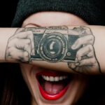 Фото татуировки с реалистичным изображением камеры на предплечье девушки. Черно-белый рисунок с детальной проработкой и ярким акцентом на красных губах.