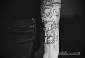 Фото татуировки в стиле line art, изображающей чертеж камеры на руке мужчины. Детализированный рисунок с использованием только черных линий.