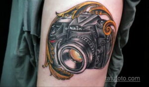 Фото тату фотоаппарата Nikon, украшенного золотыми листьями на руке мужчины. Яркий и детализированный рисунок татуировки.