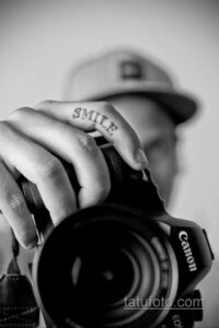 Фото тату надписи 'Smile' на пальце руки с изображением фотоаппарата. Черно-белый рисунок, каллиграфический шрифт.