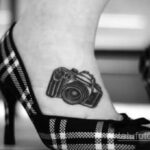 Фото тату: небольшой черно-белый рисунок фотоаппарата на лодыжке девушки. Изящная и минималистичная татуировка.