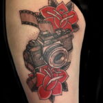 Фото тату винтажного фотоаппарата Nikon, обвитого красными розами и лентой фотопленки на бедре. Детализированный рисунок татуировки в стиле традишнл.