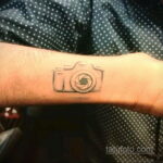 Фото тату минималистичного контурного рисунка фотоаппарата на запястье. Простой и элегантный дизайн татуировки в черном цвете.