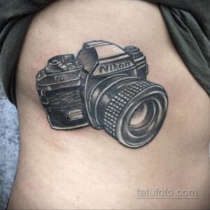 Черно-белая татуировка на плече с изображением фотоаппарата Nikon. Детализированный рисунок для поклонников бренда.