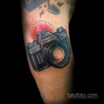 Цветная татуировка на руке с изображением фотоаппарата Canon. Детализированный рисунок с красным свечением вокруг объектива.
