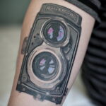 Фото тату: реалистичная черно-серая татуировка старинного фотоаппарата Rolleicord на икре. Детализированный рисунок, тени, реалистичная текстура.
