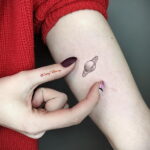 Фото тату на пальце с изображением планеты Сатурн и ее колец, выполненное в минималистичном стиле с использованием тонких черных линий.