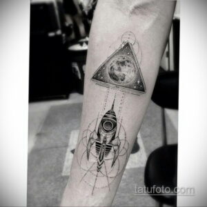 Черно-белое фото тату на предплечье с изображением ракеты внутри треугольника, с луной и звездами. Детализированный рисунок татуировки в геометрическом стиле.