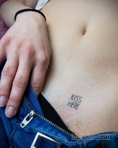Фото тату: надпись "KISS HERE" на животе. Минималистичная и провокационная татуировка, выполненная черными чернилами.