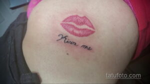 Фото тату на плече: реалистичные красные губы в поцелуе с надписью 'Kiss me' выполненной каллиграфическим шрифтом.