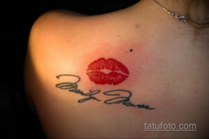 Фото тату: Красный отпечаток губ и подпись 'Marilyn Monroe' на плече женщины. Изящный и запоминающийся рисунок татуировки.