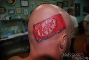 Фото татуировки KitKat на лысой голове. Реалистичное изображение шоколадного батончика с яркими цветами.