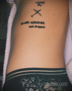 Фото тату на боку, изображающее самолет, компас и надпись 'die with memories, not dreams'. Черно-белый рисунок татуировки символизирует любовь к путешествиям и приключениям.