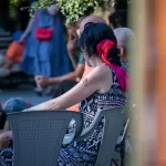 Фото тату на плече женщины: небольшой рисунок, возможно, птица или цветок. Женщина сидит на улице.