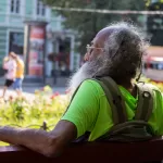 Фотография пожилого мужчины с седой бородой и татуировкой на руке, сидящего на скамейке в парке. Он смотрит в сторону.