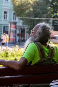 Фотография пожилого мужчины с седой бородой и татуировкой на руке, сидящего на скамейке в парке. Он смотрит в сторону.