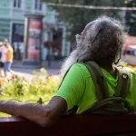 Фотография пожилого мужчины с седой бородой и татуировкой на руке, сидящего на скамейке в парке. Он смотрит в сторону.