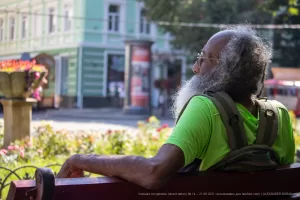 Фотография пожилого мужчины с длинной седой бородой, сидящего на скамейке в парке. На его руке видна татуировка.