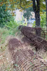 Фотография ржавой металлической арматуры, лежащей в траве рядом с деревьями и городскими постройками. Арматура выглядит старой и заброшенной.