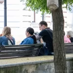 Фотография группы людей, сидящих на скамейке в парке. У одного из мужчин видна татуировка на руке, выполненная в черно-белых тонах.