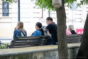Фотография группы людей, сидящих на скамейке в парке. У одного из мужчин видна татуировка на руке, выполненная в черно-белых тонах.