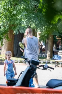Фото мужчины с рукавом татуировок, разговаривающего с маленькой девочкой в парке. Видны татуировки на руках и шее.