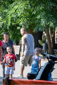 Фото тату на руках и шее мужчины, стоящего с детьми. Видны татуировки в черном цвете. Мужчина держит ребенка на руках.