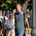 Фотография мужчины с татуировкой на руке, разговаривающего по телефону на улице. Татуировка выполнена в темных тонах и имеет сложный узор.