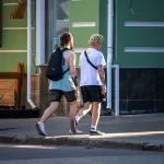 Фотография двух мужчин, один из которых с татуировкой на плече, прогуливающихся по улице в летний день.