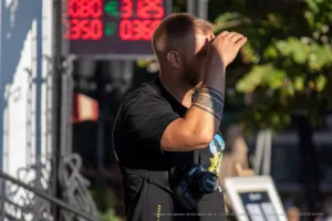 Мужчина с бородой, одетый в черную футболку, держит фотоаппарат и смотрит в видоискатель. На руке видна татуировка в стиле графики.