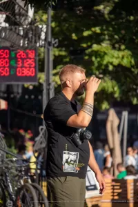 Мужчина с бородой, одетый в черную футболку, держит фотоаппарат и смотрит в видоискатель. На руке видна татуировка в стиле графики.