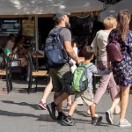 Фото мужчины с татуировкой на плече, идущего по улице с рюкзаком. Видна часть татуировки с абстрактными узорами и цветами.