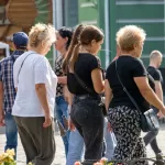 Фото женщины с татуировкой на шее, идущей по улице с двумя спутницами. Видна часть татуировки с загадочными символами.