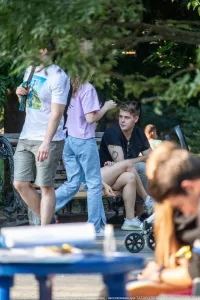 Фотография молодого человека с татуировкой на руке, сидящего на скамейке в парке. На заднем плане видны другие люди и деревья.