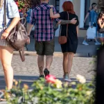 Фото тату на плече мужчины, выполненное в черном цвете. Видны контуры рисунка, татуировка частично скрыта одеждой. На заднем плане пара гуляет по улице.