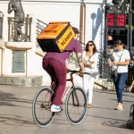 Фотография курьера на велосипеде с татуировкой на руке и оранжевой сумкой Raketa. Татуировка выполнена в темных тонах и изображает череп.
