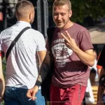 Фотография мужчины с татуировкой на руке, одетого в футболку и джинсы. На заднем плане видны другие люди и городская среда.